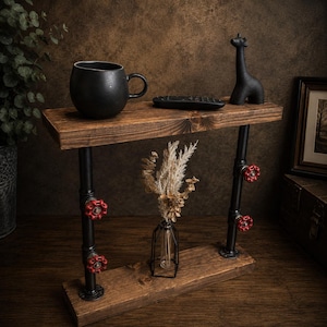 May include: A rustic, two-tiered side table with a dark wood finish. Black pipes with red valve handles support the shelves. A black mug, remote, and giraffe figurine sit on the top shelf. A vase with dried flowers is on the lower shelf.