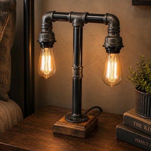 Peut inclure: Lampe de table de style industriel avec une structure en tuyaux noirs et deux ampoules de style Edison. La lampe est posée sur une base en bois et placée sur une table de chevet en bois foncé. Les ampoules mesurent environ 15 cm de long.