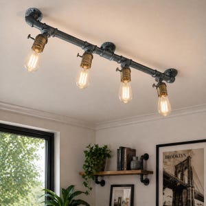 Puede incluir: Lámpara de techo de estilo industrial con cuatro bombillas estilo Edison. La lámpara está hecha de tuberías y accesorios de metal gris oscuro, con casquillos de color latón. Las bombillas están encendidas, proyectando un brillo cálido. Montada en un techo blanco.