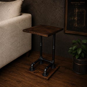 May include: A dark wood side table with a square top, supported by black metal pipes. The table is 24 inches high and 16 inches wide, with a base that rests on the floor. The design has an industrial aesthetic.