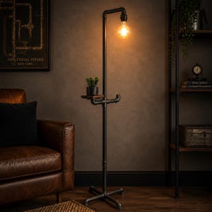 May include: Industrial-style black metal floor lamp with a small wooden shelf and a decorative lightbulb. The lamp has a cross-shaped base and a small potted plant on the shelf. The lamp is in a room with a brown leather armchair and a dark wall.