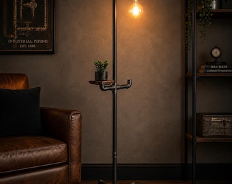 May include: Industrial-style black metal floor lamp with a small wooden shelf and a decorative lightbulb. The lamp has a cross-shaped base and a small potted plant on the shelf. The lamp is in a room with a brown leather armchair and a dark wall.