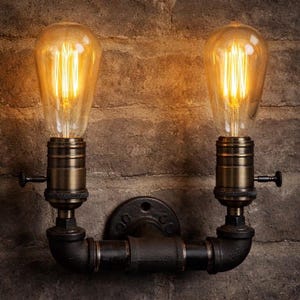 Puede incluir: Una lámpara de pared con dos bombillas de estilo Edison. La lámpara presenta un diseño de tubería negra con casquillos de lámpara de color latón y una placa de montaje circular. Las bombillas están encendidas, emitiendo un brillo amarillo cálido.