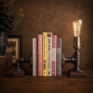 Op de afbeelding: Een industriële boekensteun met een vintage Edison-lamp. De boekensteun is gemaakt van zwarte buizen en staat op een houten voet. Boeken met titels als "Always Smiling" en "Mary Berry Fast Cakes" worden op hun plaats gehouden.
