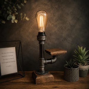 Può includere: Lampada da tavolo in stile industriale con una lampadina Edison vintage. La lampada presenta un design a tubo nero con una base in legno e un piccolo ripiano. La luce calda della lampadina crea un'atmosfera accogliente. Un pezzo decorativo.