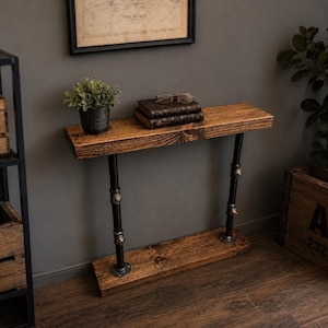 May include: A rustic, wooden console table with a dark stain. The table features black metal pipe legs and decorative fittings. A small potted plant, two books, and eyeglasses rest on the tabletop. The table is approximately 48 inches long.