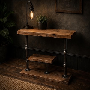 Può includere: Tavolino laterale in stile industriale rustico con finitura in legno scuro. Il tavolo presenta un ripiano superiore, un ripiano inferiore e una base, sostenuti da tubi metallici scuri. Una lampada a bulbo Edison e una pianta in vaso sono sul ripiano superiore.