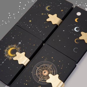 May include: Four black notebooks with celestial designs, including moons, stars, and constellations in gold and white. Each notebook has a gold star-shaped bookmark. The notebooks are arranged at an angle.