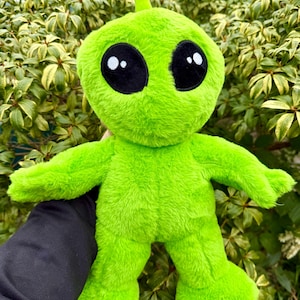 Puede incluir: Un peluche verde con forma de alienígena con grandes ojos negros y una pequeña antena. El juguete de peluche tiene una textura suave y está colocado sobre un fondo de follaje verde. El alienígena tiene los brazos extendidos y una expresión amigable.