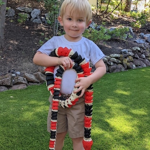May include: A young child holds a red, black, and white toy snake. The snake is made of plastic and has a realistic design.