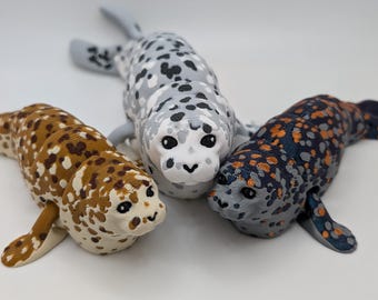 May include: Three toy seals in a row, each with a unique spotted pattern. One is brown and white, another is gray with black spots, and the third is navy blue with orange and gray spots. The seals are lying down.