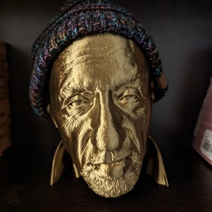 May include: A gold-colored bust of a man wearing a multicolored knit cap. The bust has detailed facial features, including a beard and wrinkles. The sculpture is displayed on a dark surface.