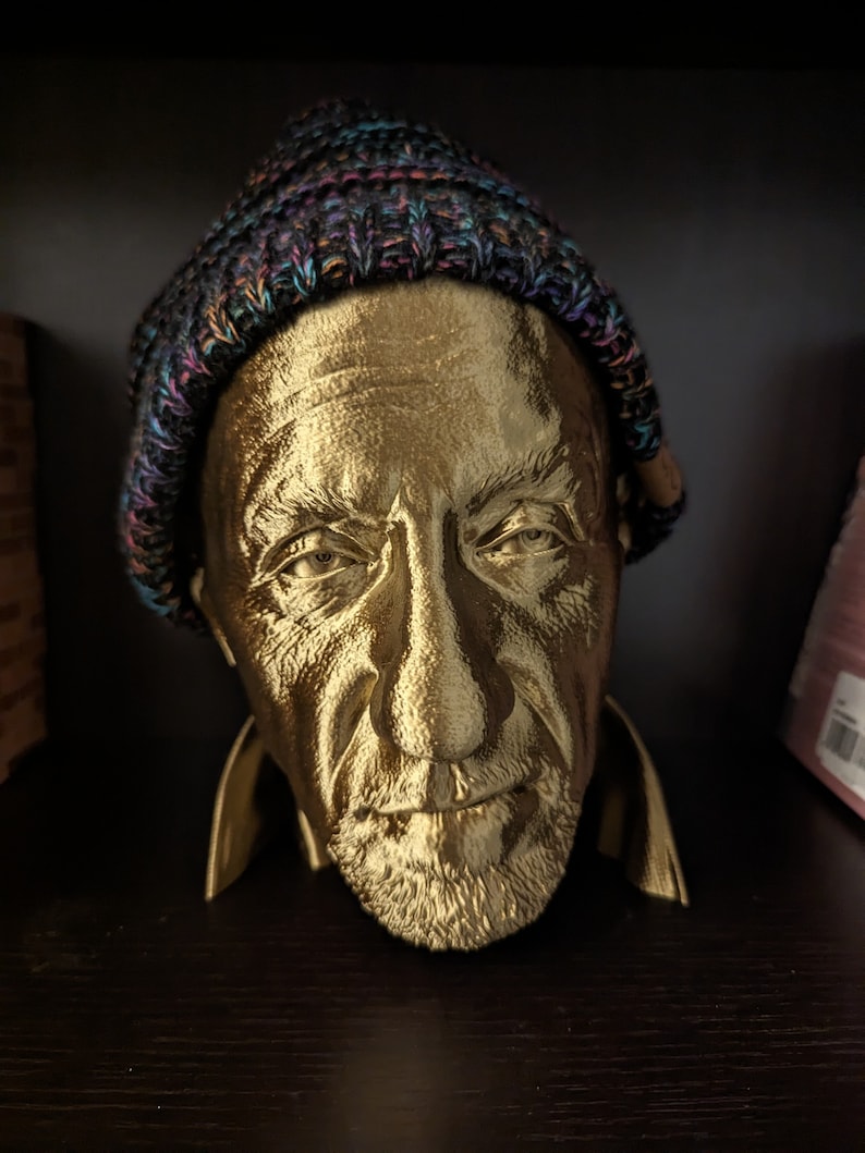 May include: A gold-colored bust of a man wearing a multicolored knit cap. The bust has detailed facial features, including a beard and wrinkles. The sculpture is displayed on a dark surface.