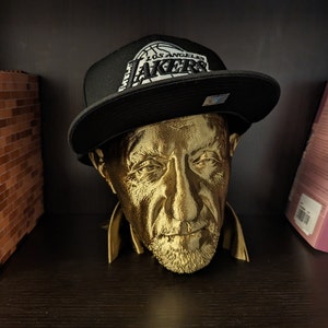 May include: A gold-colored bust of a man wearing a black Los Angeles Lakers hat. The hat has a white logo and text. The bust is on a dark shelf with books and a miniature building in the background.