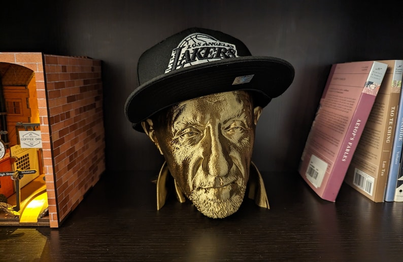 May include: A gold-colored bust of a man wearing a black Los Angeles Lakers hat. The hat has a white logo and text. The bust is on a dark shelf with books and a miniature building in the background.