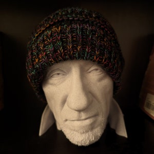 May include: A knitted beanie hat with a multicolored pattern of orange, green, and purple threads. The hat is displayed on a white bust of a person with a short beard and a collared shirt.