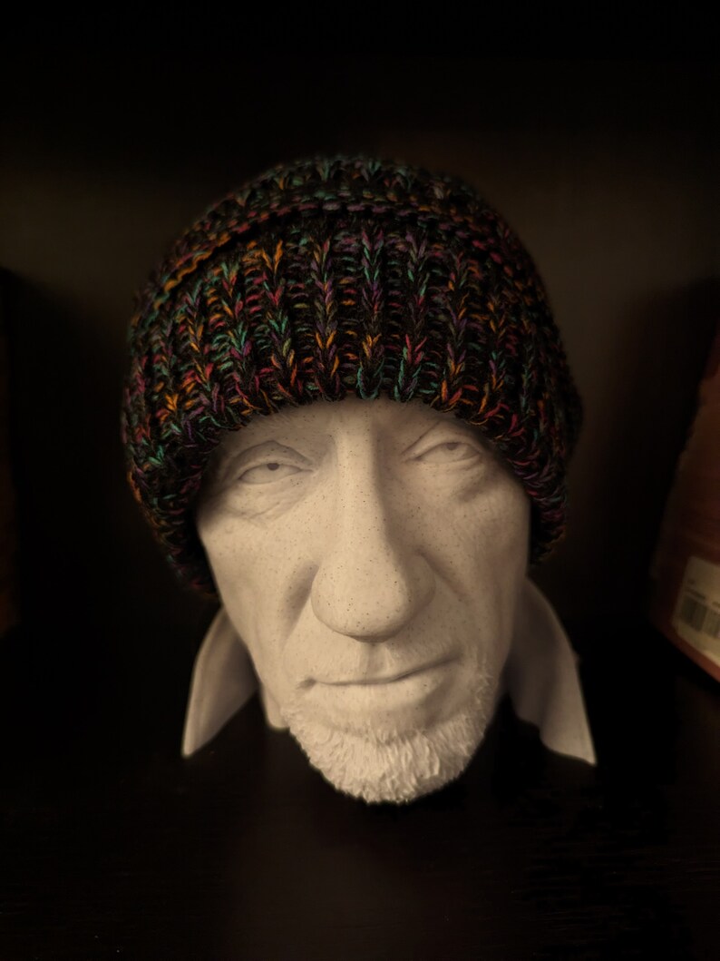 May include: A knitted beanie hat with a multicolored pattern of orange, green, and purple threads. The hat is displayed on a white bust of a person with a short beard and a collared shirt.