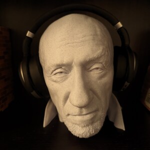 May include: A white plaster bust of a man with a short beard, wearing black over-ear headphones. The bust has a detailed face with visible wrinkles and a neutral expression. The headphones are positioned over the head, resting on the ears.