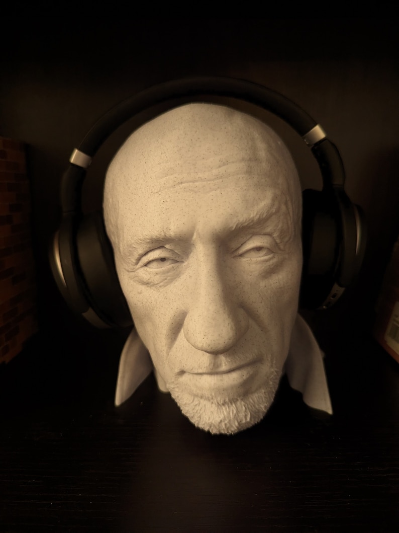 May include: A white plaster bust of a man with a short beard, wearing black over-ear headphones. The bust has a detailed face with visible wrinkles and a neutral expression. The headphones are positioned over the head, resting on the ears.