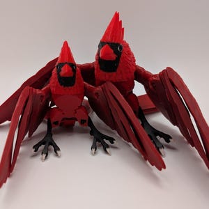 May include: Two red cardinal bird figurines with black eyes and beaks. The figurines have outstretched wings and are posed as if they are flying.