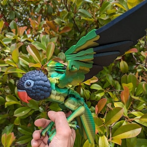 May include: A 3D printed parrot toy with articulated wings and legs. The parrot is green, black, and yellow with a black head and a red beak.