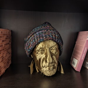 May include: A gold-colored bust of a man wearing a multicolored knit beanie. The bust is on a dark shelf, with a miniature diorama of a street scene and a row of books visible in the background. The books include titles like "Aesop's Fables."