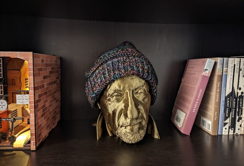 May include: A gold-colored bust of a man wearing a multicolored knit beanie. The bust is on a dark shelf, with a miniature diorama of a street scene and a row of books visible in the background. The books include titles like "Aesop's Fables."