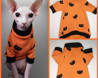 May include: Orange cat sweater with black cat faces printed on it. The sweater has black trim around the neck and sleeves. A hairless cat is wearing the sweater.