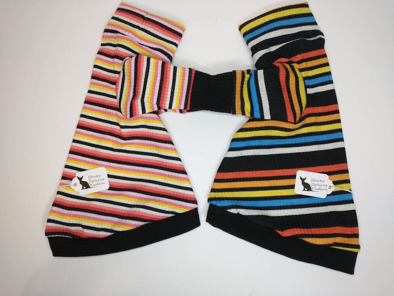 May include: Two striped dog sweaters made from black ribbed fabric. One sweater has pink, yellow and white stripes. The other sweater has blue, orange and white stripes. Both sweaters have black ribbed cuffs and collars. The sweaters have tags that say "Skinny Sphynx Fashion".
