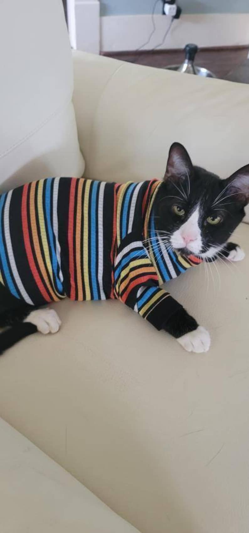 May include: A black and white cat wearing a colourful striped shirt, lying on a white sofa.
