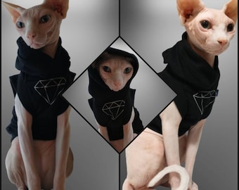 May include: Three Sphynx cats wearing black hooded shirts with a diamond graphic. The shirts have a white diamond design on the front.