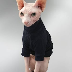 May include: A Sphynx cat wearing a dark blue turtleneck sweater. The cat has pink skin, large ears, and green eyes. The sweater covers the cat's torso and arms, providing a contrast to its hairless body. The background is a gradient of gray.