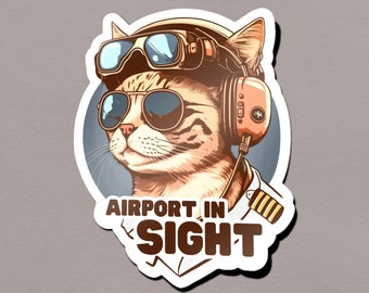 Könnte beinhalten: Ein Aufkleber mit einer Cartoon-Katze, die eine Fliegerbrille und ein Headset trägt, mit dem Text "AIRPORT IN SIGHT" darunter. Die Katze trägt eine Pilotenuniform mit goldenen Streifen.