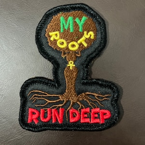 Può includere: Un cerotto ricamato nero e marrone con un disegno di albero. L'albero ha un tronco e radici marroni, e un cerchio verde e giallo in alto con il testo "MY ROOTS" in giallo. Il testo "RUN DEEP" è ricamato in rosso nella parte inferiore del cerotto.