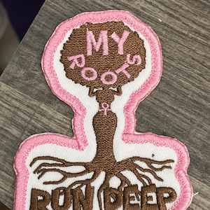 Può includere: Una toppa ricamata con un design a forma di albero in marrone e rosa. L'albero ha un afro marrone con le parole "MY ROOTS" scritte su di esso. Il tronco dell'albero è marrone e le radici sono marroni e si estendono. Le parole "RUN DEEP" sono scritte in marrone sotto le radici. La toppa è circondata da un bordo rosa.