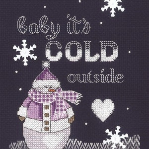May include: Cross-stitch design on a dark blue background. A snowman wearing a purple scarf and hat is featured, along with snowflakes and the phrase "baby it's COLD outside" in white stitching. A white heart is also included.
