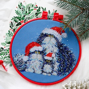 May include: A cross-stitch embroidery of three cats wearing Santa hats, set in a red hoop against a blue background. The cats are white and blue, with yellow eyes. The background includes a sprig of evergreen.