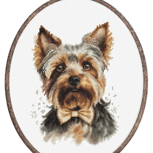 May include: A cross-stitch portrait of a Yorkshire Terrier dog wearing a bow tie, framed in an oval wooden hoop. The dog's fur is detailed in brown, black, and tan. The background is white, with a decorative wooden frame.