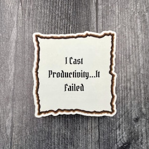 May include: A white sticker with a brown border and the text "I Cast Productivity...It Failed" in a gothic font. The sticker is on a gray wooden surface.