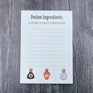 May include: A white notepad with the text "Potion Ingredients" and the words "COFFEE | CHAOS | DEADLINES" printed at the top. The notepad has lined pages and three potion bottle illustrations at the bottom. The notepad is on a wooden surface.