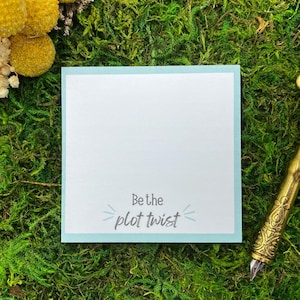 May include: A square, light blue-bordered notepad with the text "Be the plot twist" sits on a bed of green moss. A decorative gold pen and a feather are to the right. Yellow and white dried flowers are in the upper left.