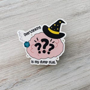 May include: A sticker featuring a pink brain with question marks, a wizard hat, and the text "Overthinking is my dump stat." The hat is black with gold stars and a yellow band. A blue and teal die is also present.