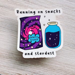 May include: A sticker featuring a purple and pink snack bag labeled "Galactic Crunch" and a blue jar with a starry liquid. The text "Running on snacks and stardust" is above and below the images.