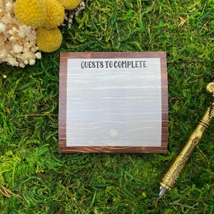 May include: A square notepad with a wood grain border and the text "QUESTS TO COMPLETE" printed at the top. The center of the notepad features a light gray area with a subtle wood grain pattern, providing a space for writing tasks or notes.