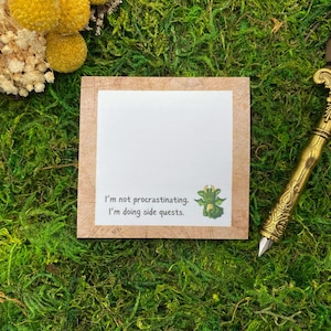 May include: A square notepad with a light brown border and a white center featuring the text I'm not procrastinating. I'm doing side quests. and a small green dragon illustration, accompanied by a metallic gold pen with an ornate engraved pattern.