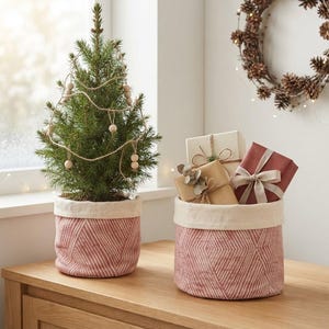 Op de afbeelding: Een kleine groene naaldboom versierd met een slinger van houten kralen staat in een roze en wit patroon stoffen mand. Daarnaast staat een bijpassende stoffen mand met drie ingepakte cadeaudoosjes, vastgebonden met linten en touw.