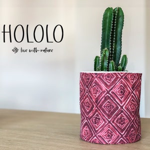 Puede incluir: Una planta de cactus en una maceta de tela roja y blanca con estampado. La maceta tiene un diseño geométrico con pequeñas formas de flores. El texto "HOLOLO live with nature" está en la pared detrás de la maceta.