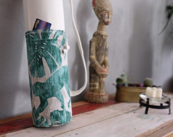 May include: A white yoga mat carrier with a green and white tropical leaf print. The carrier has a drawstring closure and a shoulder strap. A credit card is visible in the pocket.