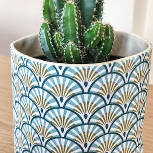 Könnte beinhalten: Dekorativer Blumentopf mit einem geometrischen Fächermuster in Türkis, Weiß und Gold. Der Topf enthält einen grünen Kaktus mit scharfen Stacheln. Der Topf hat einen neutralfarbenen Innenraum und steht auf einer Holzoberfläche.