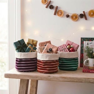 May include: Three fabric storage baskets with white cuffs and horizontal stripes in dark purple, rust orange, and forest green. Each basket is filled with wrapped gift boxes featuring velvet ribbons, pinecones, and ornaments.